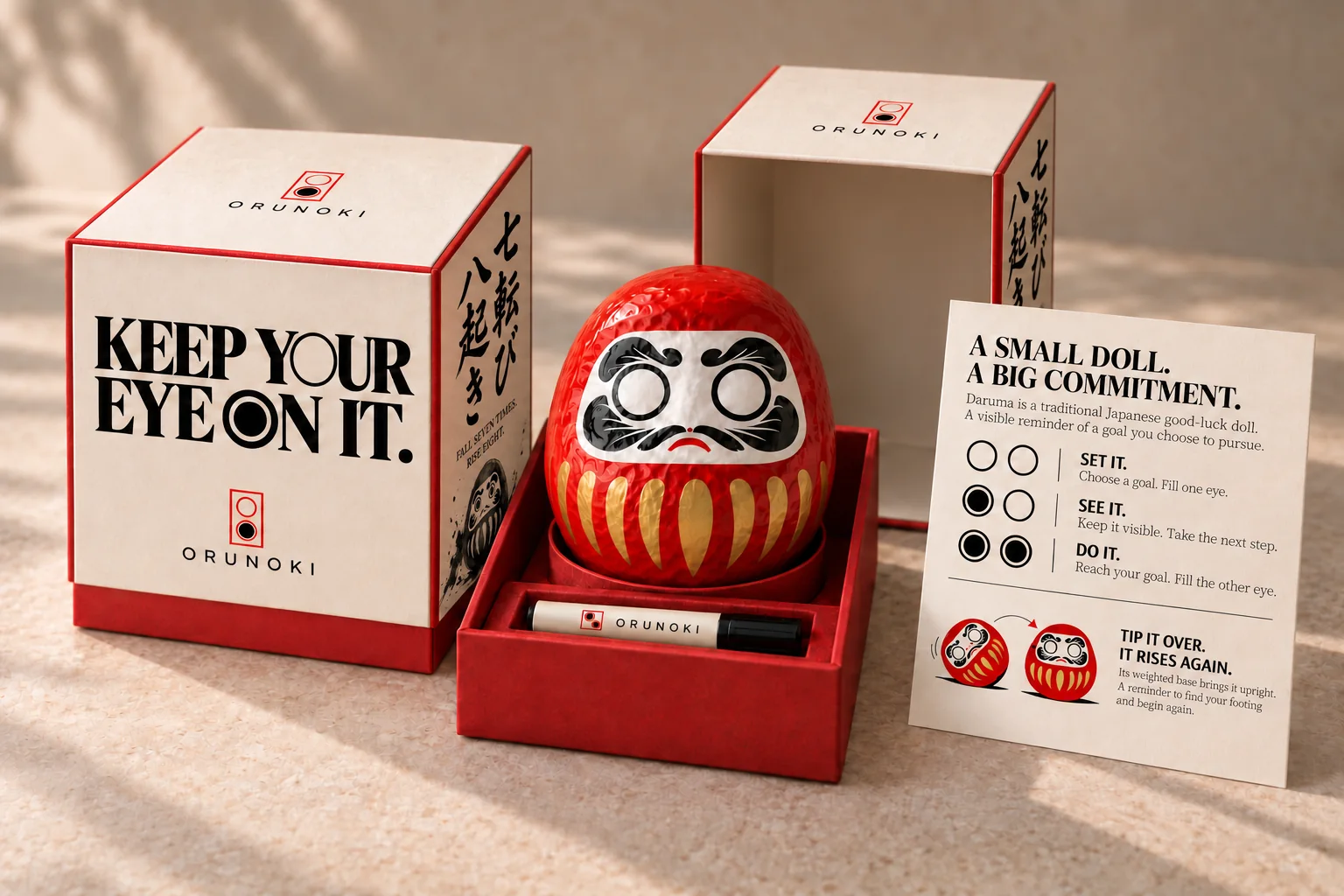 An open gift box holding a red Daruma with blank eyes and a marker pen, next to the closed box and a card with the ritual.